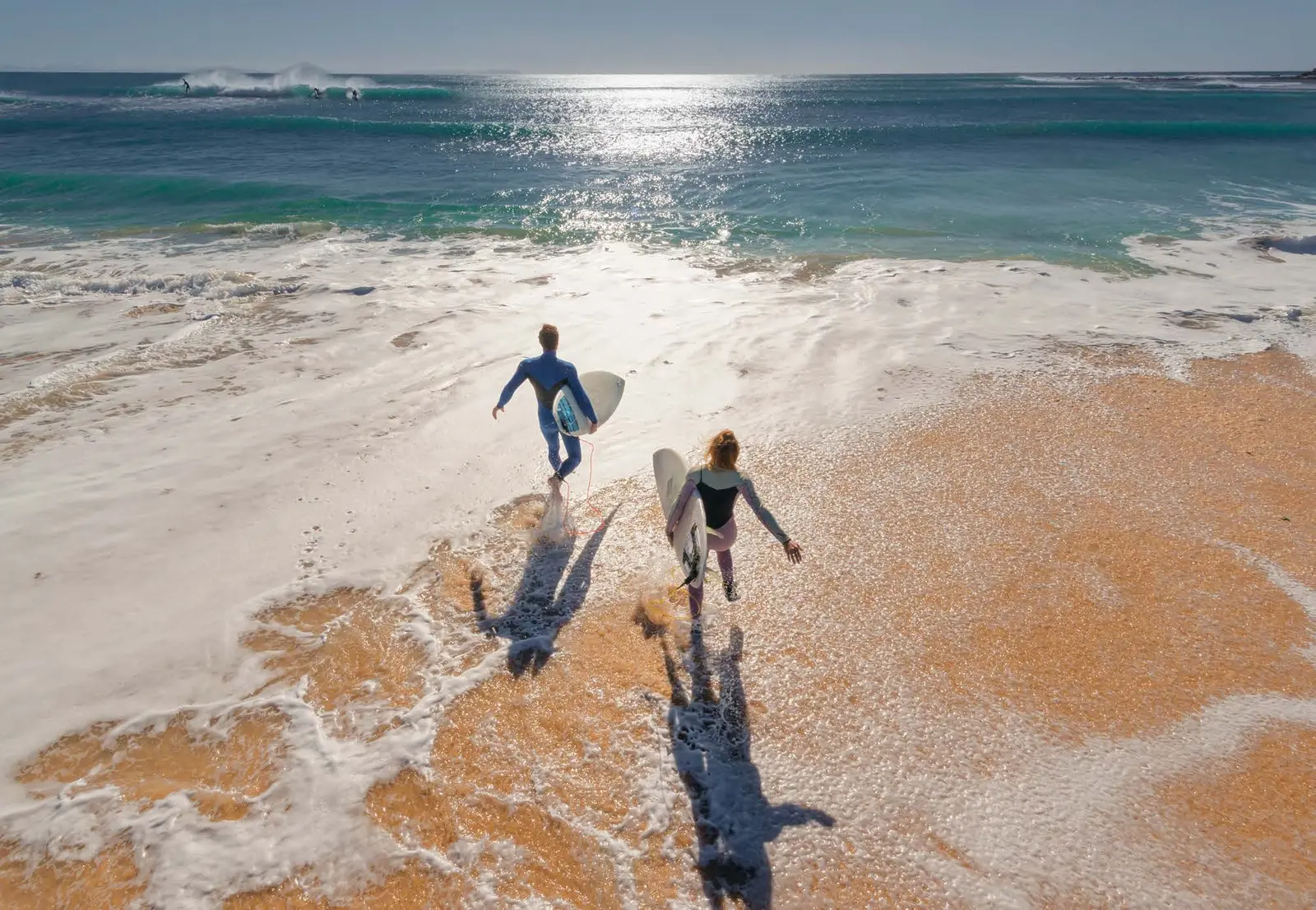 Surfers heading into rolling coastal waves directly along pristine beaches surrounding Capeside Marina Residences Sobha Siniya Island.